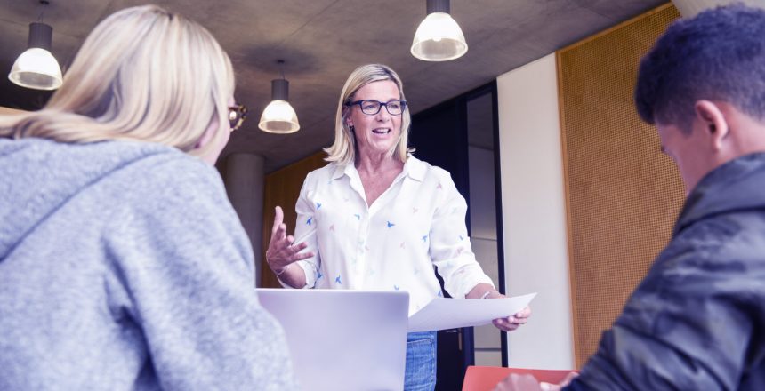 Mature woman gesturing and holding paper, male and female college students listening in forground