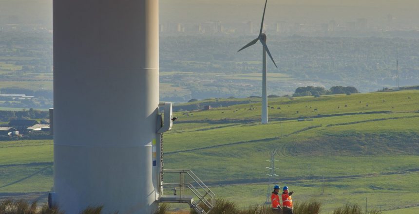two wind farm engineers walk away from the door way of a wind turbine. they are wearing orange hi vis jackets and blue hard hats . one is male , one is female. In the background wind turbines can be seen across the landscape down to a distant city .