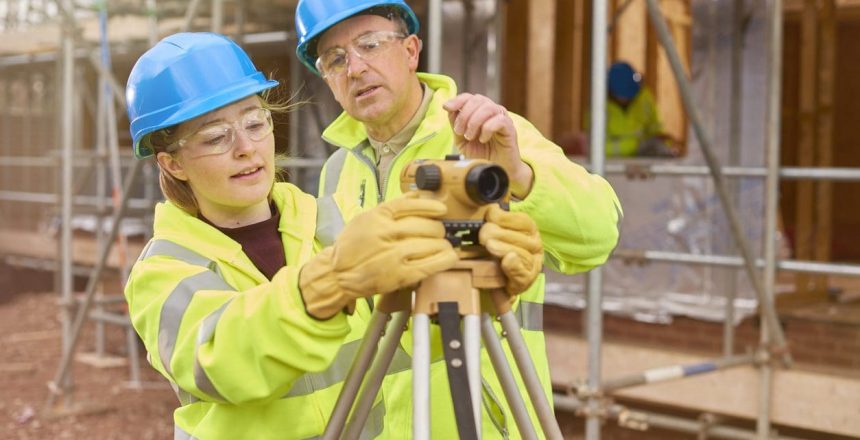 a female construction worker stands behind a builder's level on a  building site .Behind her a co-worker walks across the development .